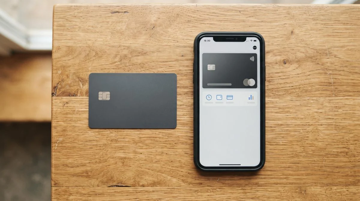 Vista superior de una tarjeta de crédito física junto a un teléfono inteligente que muestra una billetera digital abierta, sobre una mesa de madera